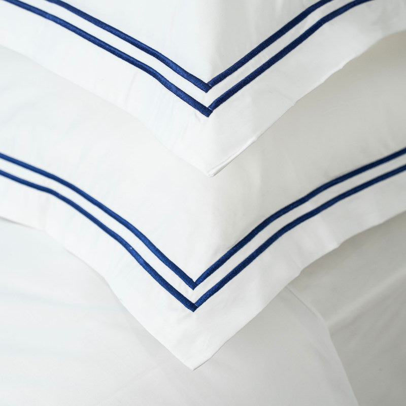 A white cotton sateen flat sheet with navy blue cord trim on the top hem, part of a bed linen set.