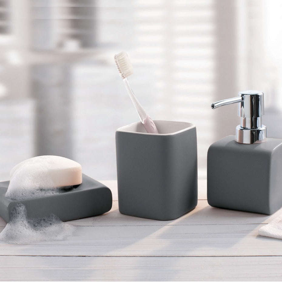 Three-piece gray bathroom vanity set including a toothbrush holder with a toothbrush, a soap dispenser with a chrome pump, and a soap dish with a bar of soap, arranged on a light wood surface with a blurred bathroom background.