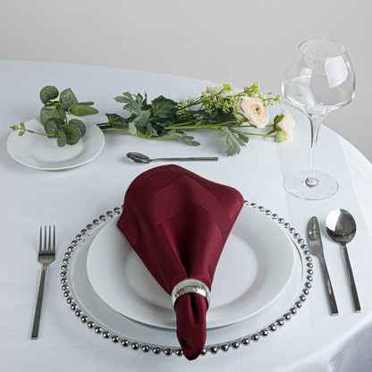 A styled table setting featuring a deep burgundy folded napkin with a silver ring, layered over white plates with a silver beaded charger plate. The setting includes silver cutlery, a clear wine glass, a small dish of green leaves, and a floral centerpiece on a white tablecloth.