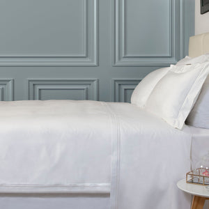 A white flat sheet on a bed with a decorative trim on the top edge, displayed in a bedroom setting with a nightstand and lamp.