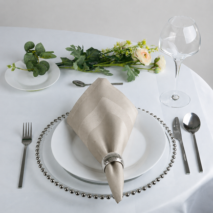A beige striped napkin folded into a cone and secured with a silver ring, displayed on a white plate at a place setting on a white tablecloth, with a wine glass, cutlery, and flowers in the background.