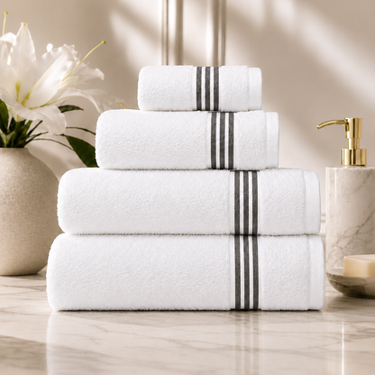 A stack of four white towels with two black stripes on the center of each towel, arranged on a marble countertop with a vase of white lilies and a soap dispenser in the background.