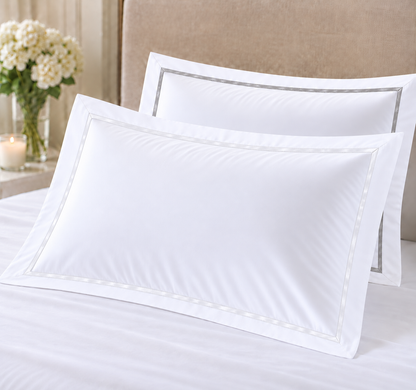 Two white pillowcases with delicate silver-gray piping borders displayed on a bed with white linens, set against a neutral beige headboard and a side table with flowers.