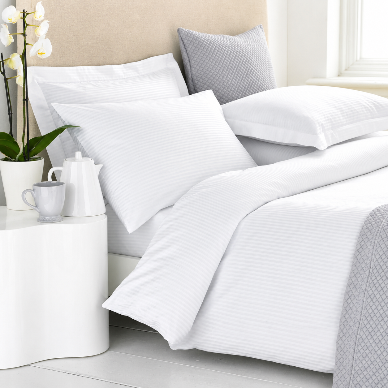 White striped duvet cover and pillowcases on a bed, styled with a gray textured pillow, white bedside table, and orchid flowers.