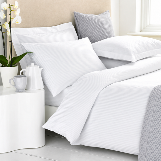 White striped duvet cover and pillowcases on a bed, styled with a gray textured pillow, white bedside table, and orchid flowers.