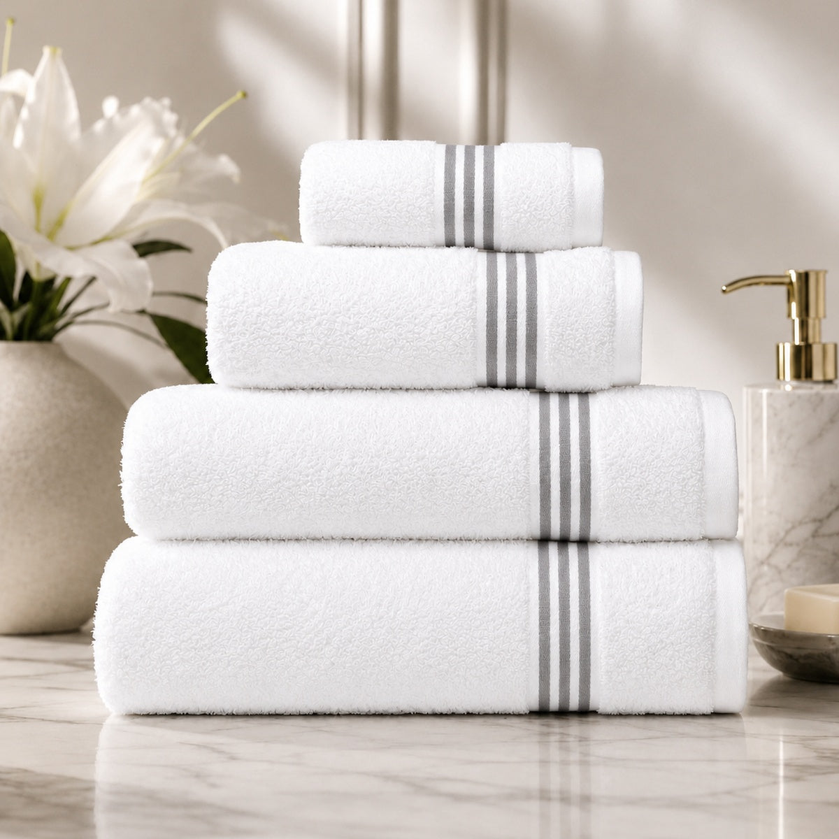 A stack of four white cotton towels with two gray stripes on the long ends, arranged on a marble surface with a vase of white flowers and a soap dispenser in the background.