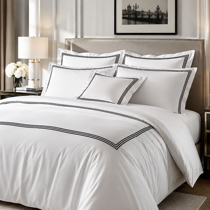 A neatly made bed in a styled bedroom setting, featuring a white duvet cover and multiple matching pillows with thin black piping trim. The room includes a beige upholstered headboard, bedside tables, a lamp, curtains, and framed wall art.