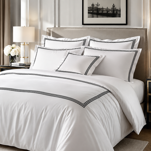 A neatly made bed in a styled bedroom setting, featuring a white duvet cover and multiple matching pillows with thin black piping trim. The room includes a beige upholstered headboard, bedside tables, a lamp, curtains, and framed wall art.
