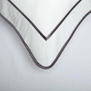 Close-up of a white pillowcase corner with dark grey ribbon borders and a rounded piped edge.