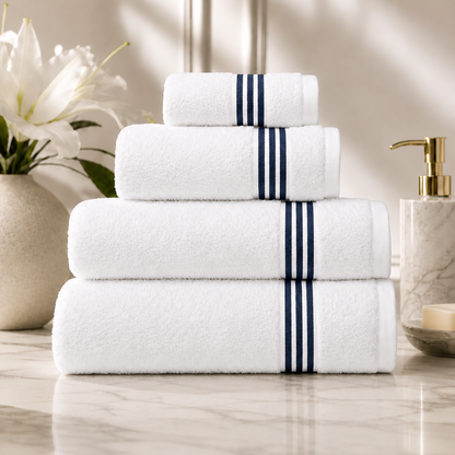 A stack of four white towels with navy blue stripe accents on the ends, arranged on a marble countertop with a vase of white lilies and a soap dispenser in the background.