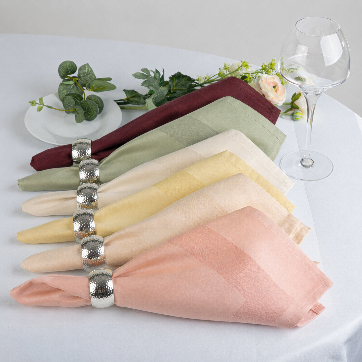 Six folded fabric napkins in dusty pink, peach, pale yellow, sage green, burgundy, and cream, each tied with a silver textured napkin ring on a white tablecloth, styled with a wine glass, flowers, and greenery.