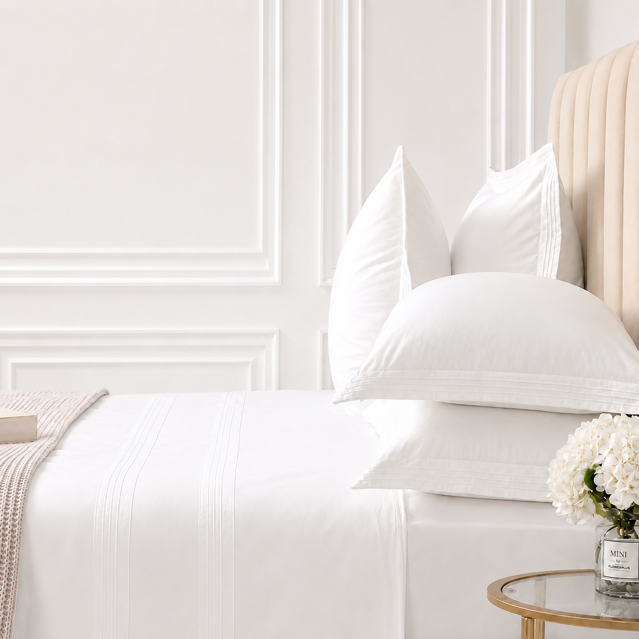 A styled bedroom scene showing a neatly made bed with white bedding, multiple white pillows, a cream upholstered headboard, white wall molding, a small round side table with flowers, and a folded knit throw at the foot of the bed.