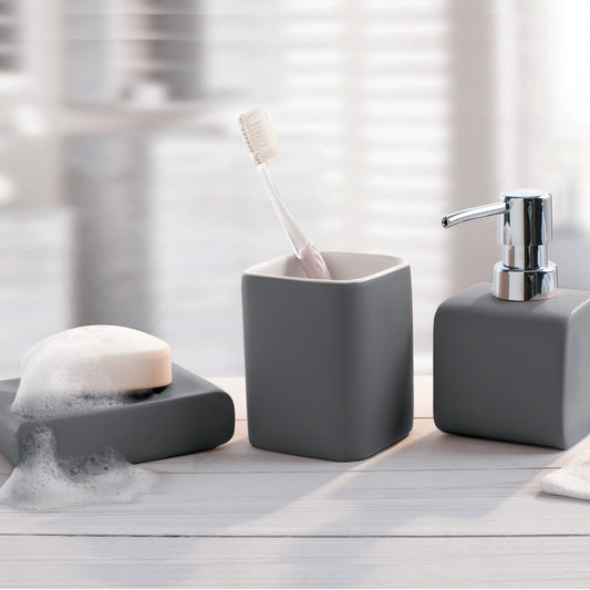 Three-piece gray bathroom vanity set including a toothbrush holder with a toothbrush, a soap dispenser with a chrome pump, and a soap dish with a bar of soap, arranged on a light wood surface with a blurred bathroom background.