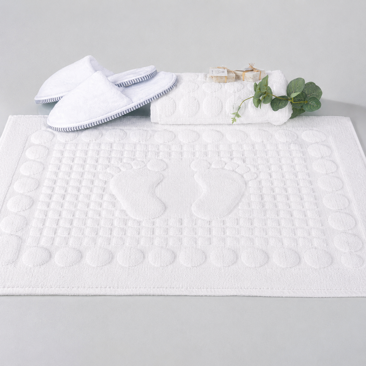 A white textured bath mat with footprints in the center, styled with white slippers, a folded towel, and a small green leaf sprig on a light gray background.