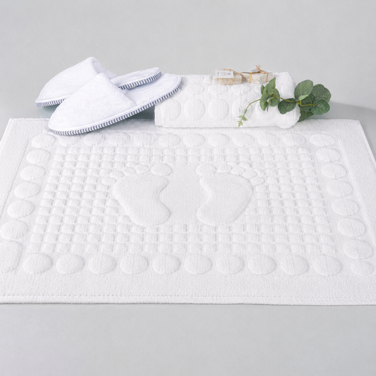 A white textured bath mat with footprints in the center, styled with white slippers, a folded towel, and a small green leaf sprig on a light gray background.