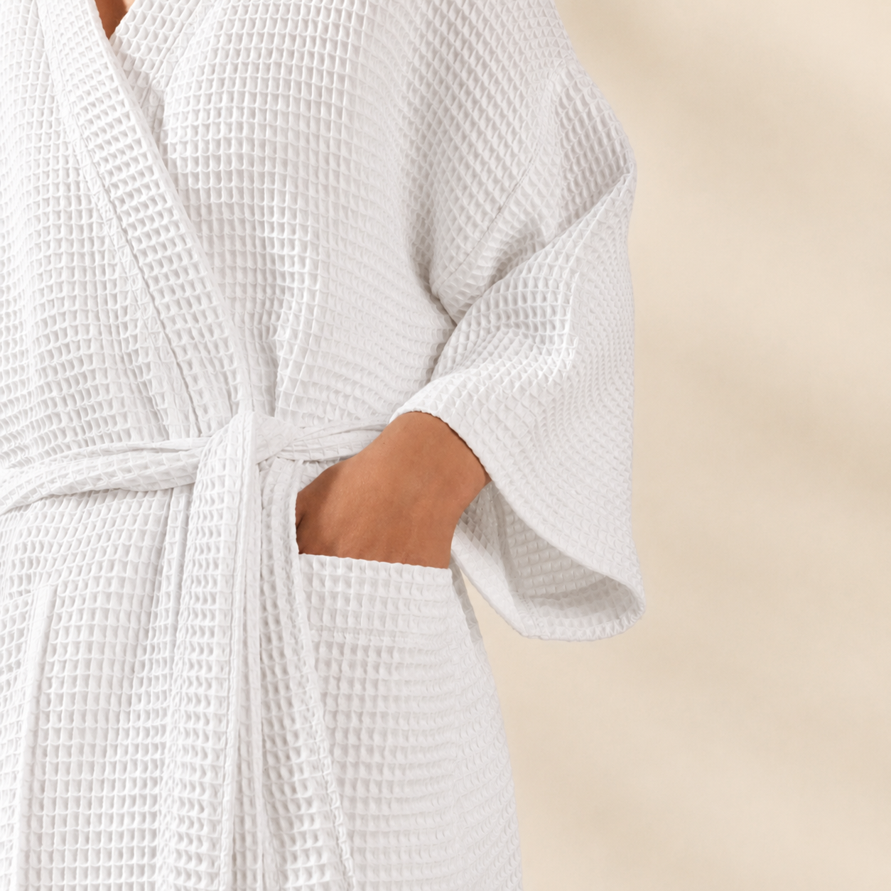 Close-up of a person wearing a white waffle-textured robe with a tie belt and side pocket, against a soft beige background.