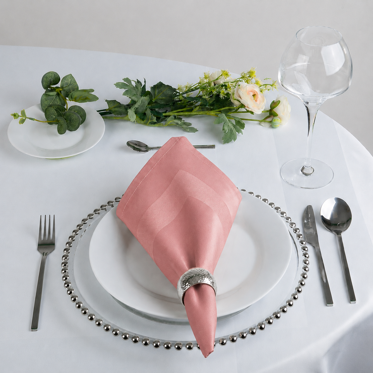 A pink satin napkin with a subtle diagonal stripe pattern, folded into a cone shape and held in a silver ring, set on a white plate on a white tablecloth. The table setting includes a silver beaded charger plate, silver cutlery, a clear wine glass, and green floral accents.
