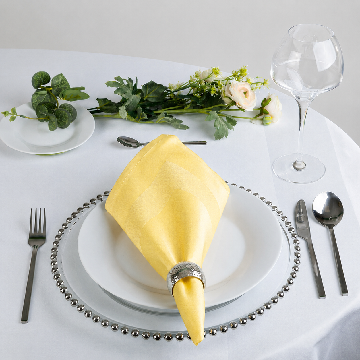 A yellow fabric napkin folded into a cone shape and secured with a silver ring, displayed on a white plate set on a round table with silver cutlery, a wine glass, and floral accents.